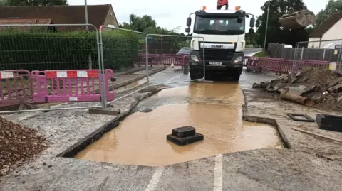 A road that has been dug up, showing a large amount of dirty brown water in it, with a lorry behind it. There are barriers, metal and plastic, and rows of rubble and mud piled up around it.