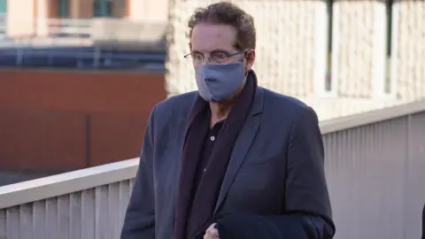 Oli Constable/BBC Chris Brain leaving court following an earlier appearance. He wears glasses and a face covering, as well as a dark-coloured blazer and scarf.