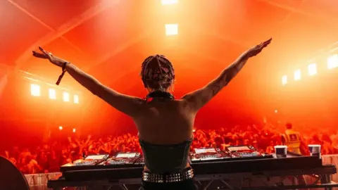 A DJ with her back to the camera holding her arms in the air as she stands over her DJ set. The lighting is orange.