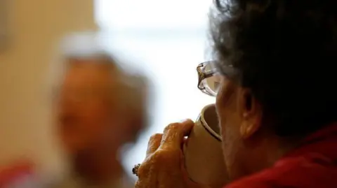 Two women: in the foreground a woman sips from a white tea cup. Her left ear, left cheek, glasses and left hand can be seen. The blurred image of the second woman is visible in the background.