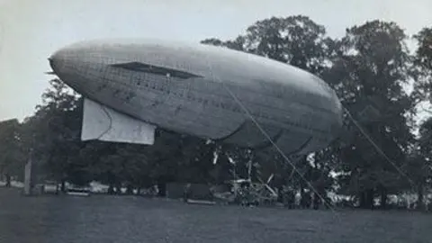 Michelle Bullivant A black and white image of a Beta II airship floating above grass at Jesus Grove Cambridge, in 1912. It is tied to the ground with ropes. Behind it is an avenue of trees.