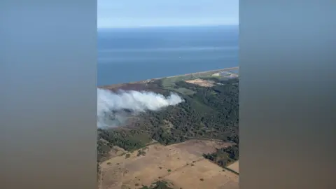 Rob Forsyth In a picture taken from the air, smoke is drifting from right to left from a wooded area. A large area of open ground is in the foreground, and the sea is at the top of the picture.
