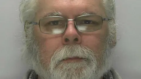 A mugshot of a man in his 60s who has a medium-length, grey beard and white hair. He is wearing rectangular, silver glasses and is looking blankly ahead at the camera.