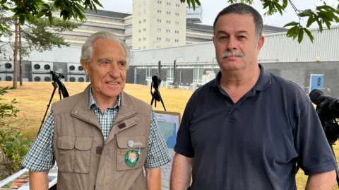Stuart Howells/BBC Peter Merchant, a grey-haired man in a check shirt and brown waistcoat, stands next to Robert James, a man with dark, greying hair and a grey moustache, wearing a navy polo shirt. Industrial buildings can be seen behind, and three monoculars on tripods