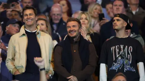 Getty Images Tom Brady, Minority Owner of Birmingham City (L) with David Beckham and Romeo. The three men stand side by side. The two older men are smiling and wear jackets. Romeo, pictured on the right, wears a T-shirt with "Texas" written on, a baseball cap backwards and a long white top.