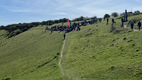 BBC/George Carden A crowd of people gathering on a green hillside. 
