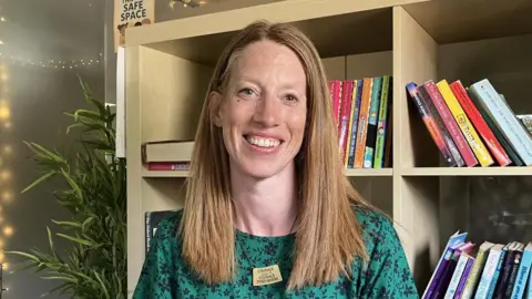 BBC/Phil Bodmer A woman with red hair and a green top looks at the camera smiling, with rows of books on shelves behind her.