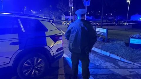 Un oficial de policía está parado de espaldas a la cámara. Es de noche. Está parado junto a un auto de emergencia. Hay una cinta acordonante del área.