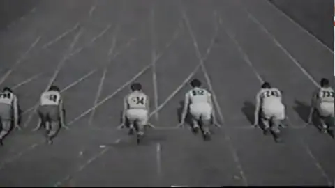 Jim Pennington A black and white photo with several athletes at the start line of a 100m track. They have their backs to the camera and are all on their knees posed to start the race 