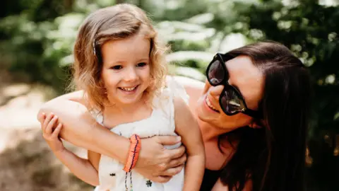 Sarah J Scott Photography Lucy with her arms round Margo, with sunglasses on, smiling. Margo has shoulder length fair hair, Lucy has long dark hair.