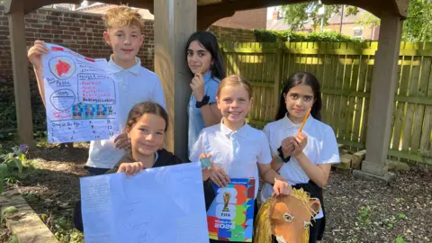 Image shows five children aged 11 wearing white polo shirts and holding things. Two of them are holding pencils, two are holding posters and one girl is holding a FIFA World Cup 2026 sticker book as well as a paper mache lion head mask.