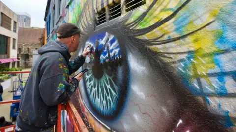 Upfest A man is spray painting a very detailed eye on to the walls of Quakers Art Lane. The iris of the eye is blue - the bottom half appears to represent the ocean and the top half appears to show the sky and black figurines.The eye has long eye-lashes and yellow paint surrounding it.