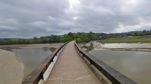 A narrow pedestrian bridge with wooden railings spans a muddy river or estuary. The sky is overcast with grey clouds, and trees and greenery line both sides of the water. No people are present in the scene.