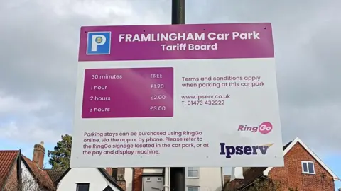 Supplied A pink and white sign attached to a black pole. It says Framlingham Car Park Tarrif Board. The cost of parking at that particular car park is also set out on the sign. Houses are also in the background of the image.