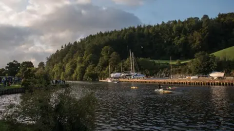 A river with a bush in the foreground and a bank with some trees and a field on the far side and a quay.