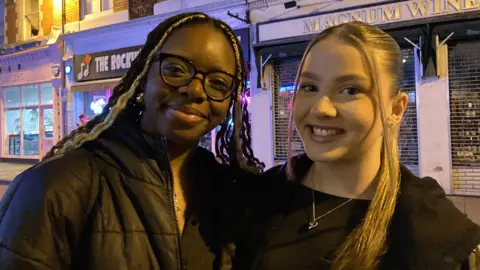 Two young women are standing next to each other on a dimly-lit street in Swindon and smiling at the camera. One is a black woman with long hair and glasses, wearing a black puffer coat. The other is a white woman also wearing all black with a silver heart necklace. A sign above them reads 'Magnum Wines'.