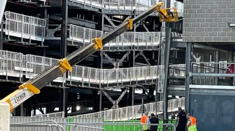 Helen Mulroy/BBC A group of people watch on as a large yellow crane is directed towards a gap in the car park where the vehicle fell from. A long metal barrier is hanging off the side of the building.