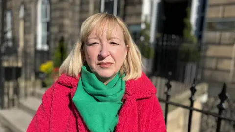 Andrea Bradley outdoors in bright sunlight wearing a red coat and green scarf, standing in front of a row of stone buildings and black railings.