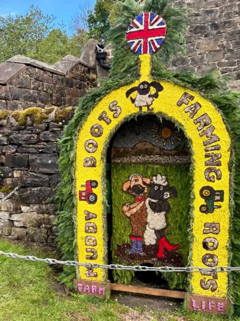 Tissington Well Dressings A small water spring below a stone wall. Above it is a bright floral display of Shaun the Sheep and a dog. The words "Muddy boots farming roots" are included in the design, as is the union flag.
