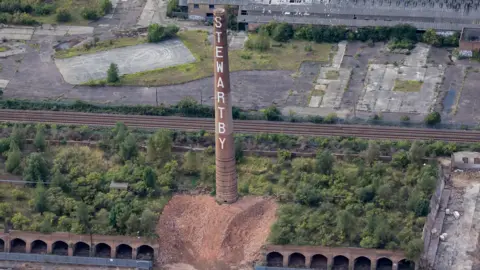 Getty Images A tall brick chimney standing alone in the centre of the image. The word "Stewartby" is painted vertically in large white letters on the chimney.