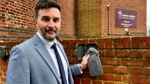 Sara Smith/BBC Shaun Meaney, wearing a light blue jacket, shirt and dark blue tie, holds up a pouch for a smartphone, in front of a brick wall, with the school's sign in the background. 