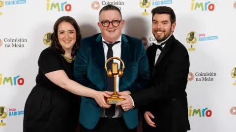 BBC John Toal in the centre wearing a green/blue velvet jacket smiles as he holds a gold microphone IMRO award. On one side a woman with brown hair in a black dress reaches over to touch the microphone, on the other side a man in a tuxedo does the same