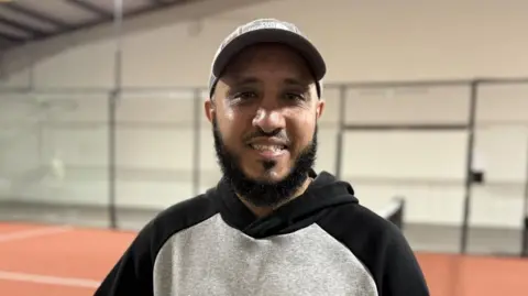 Atique smiles at the camera, posing on the padel court. He is wearing a grey hoodie and cap.