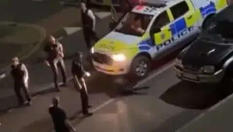 Amarjit Kharaud A number of police officers in black uniforms next to a police car. The cow is stuck underneath the police car. 