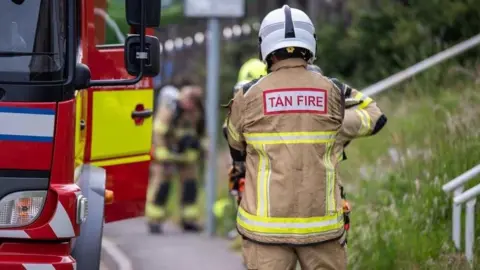 A firefighter standing beside a fire engine at what appears to be an incident scene or training exercise. In the foreground, a person is seen from behind wearing firefighting protective clothing with high-visibility reflective stripes and a white helmet. The words "Tan Fire" are visible on the back of the jacket, indicating the Welsh word for "fire". On the left side of the image is part of a red fire engine with yellow fluorescent panels and emergency-service markings.