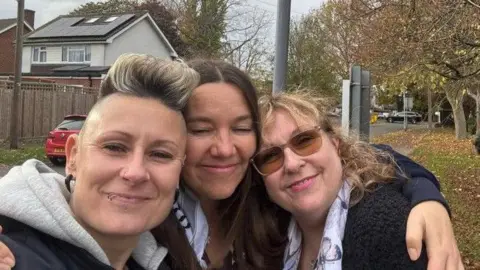 PCC Office L to R, Lauren Preston, Laura Hughes and Mary Sharp. Laura has her arms around the other two. They trio are smiling. They are wearing coats. Behind them is a street, trees and cars. 