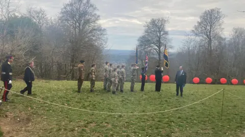 Surrey County Council A view of a group of people in army wear with flag bearers at the front and others in military uniform behind. In the distance the hill drops down to show a town and trees further off.