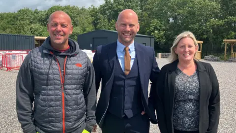 The three co-founders standing in the Academy's gravel training yard. They are looking towards the camera smiling. William King wears a padded body warmer with red zip over a dark grey hooded jumper. Daniel Gee is wearing a navy three piece suit with thin burnt orange tie. Rachel Holliday is wearing a mosaic print black and grey tope with a black suit jacket. 