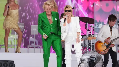 PA Media A man and a woman are on stage together, holding microphones. There are other musicians behind them. The man is wearing a green suit, while the woman is wearing a white suit with tasseled trousers.