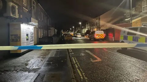 A line of police tape cordons off a residential street in York, with several police vehicles parked by the side of the road.