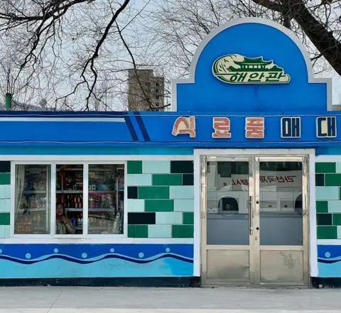 Joe Smith, a photo of the outside of a blue food store in North Korea. The exterior has green titles and there is a woman looking through a window