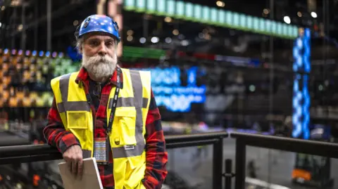 TIL BUERGY/EPA-EFE/REX/Shutterstock A man with a large beard wearing a flannel shirt, a helmet and a high-vis jacket poses for the camera as lighting infrastructure is erected behind him in a large stadium.