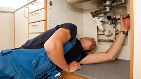 Getty Images Plumber in blue overalls and a black t-shirt under a sink with a wrench in his hand.