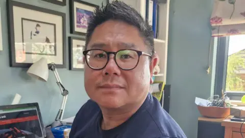 Pak Wai Hung A man with glasses sitting at a desk with a laptop open in the background. He is wearing a blue T-shirt and is sat next to a window with a number of pictures hanging on the wall.