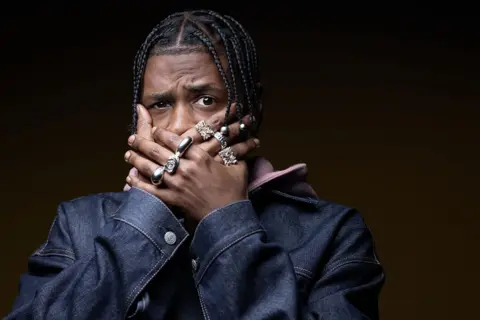 JOEL SAGET / AFP / GETTY IMAGES A man wearing a denim jacket and copious silver jewellery covers his mouth with his hands and feigns a shocked expression.