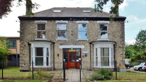 The outside of John Symons House with light bricks, white sash windows and a brown wood front door