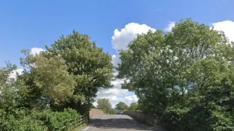 Google A road, with trees overhanging from either side, and a pavement on the left hand side.
