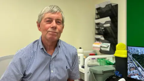 A person with short grey hair wearing a blue-and-white striped shirt sits in a clinical office setting, with shelves, trays, and medical supplies visible in the background alongside a computer monitor.