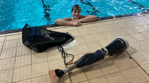 A teenage girl in a swimming pool with her arms crossed at the ledge, She is wearing goggles and a black and red swimming cap. In front of her on the pool edge is a black bag and a prosthetic leg