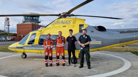 The Air Ambulance Service Four people, two in orange flying suits, one in a green ambulance uniform, stand in front of a yellow and grey helicopter at an airbase