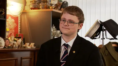 Charlie Elphinstone wears glasses, a white suit, dark tie and suit, he is surrounded by antique ornaments and hats in the conservatory of his family home