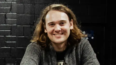 James Taylor is wearing a grey jumper. Behind him is a black wall.
