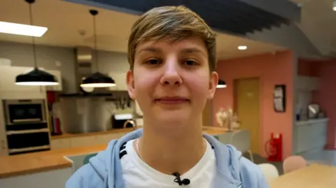 Amy McMullen standing in a kitchen area, looking directy at the camera. She has short brown hair and is wearing a white t-shirt and light blue hoodie.