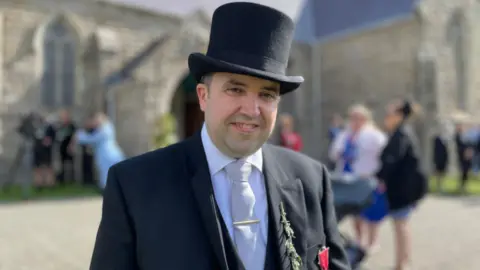 David Ashford, a man who wears a suit and top hat smiles in front of St John's Church.