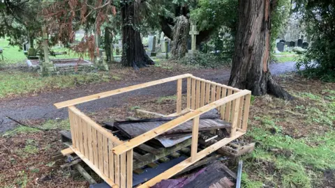 A pile of waste can be seen in the foreground, including planks of wood and a piece of pine furniture. There are grave stones a short distance from the pile of rubbish as well as some large trees.