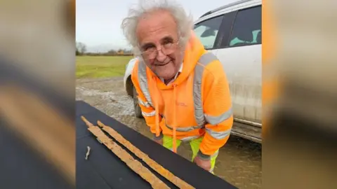 James Haris Buzz Busby with grey hair and wearing clear glasses, is in a bright orange hoody with reflective strips. He is standing next to a table with two swords on. They are long and rusty. There is a field in the background, with a white vehicle behind Buzz.